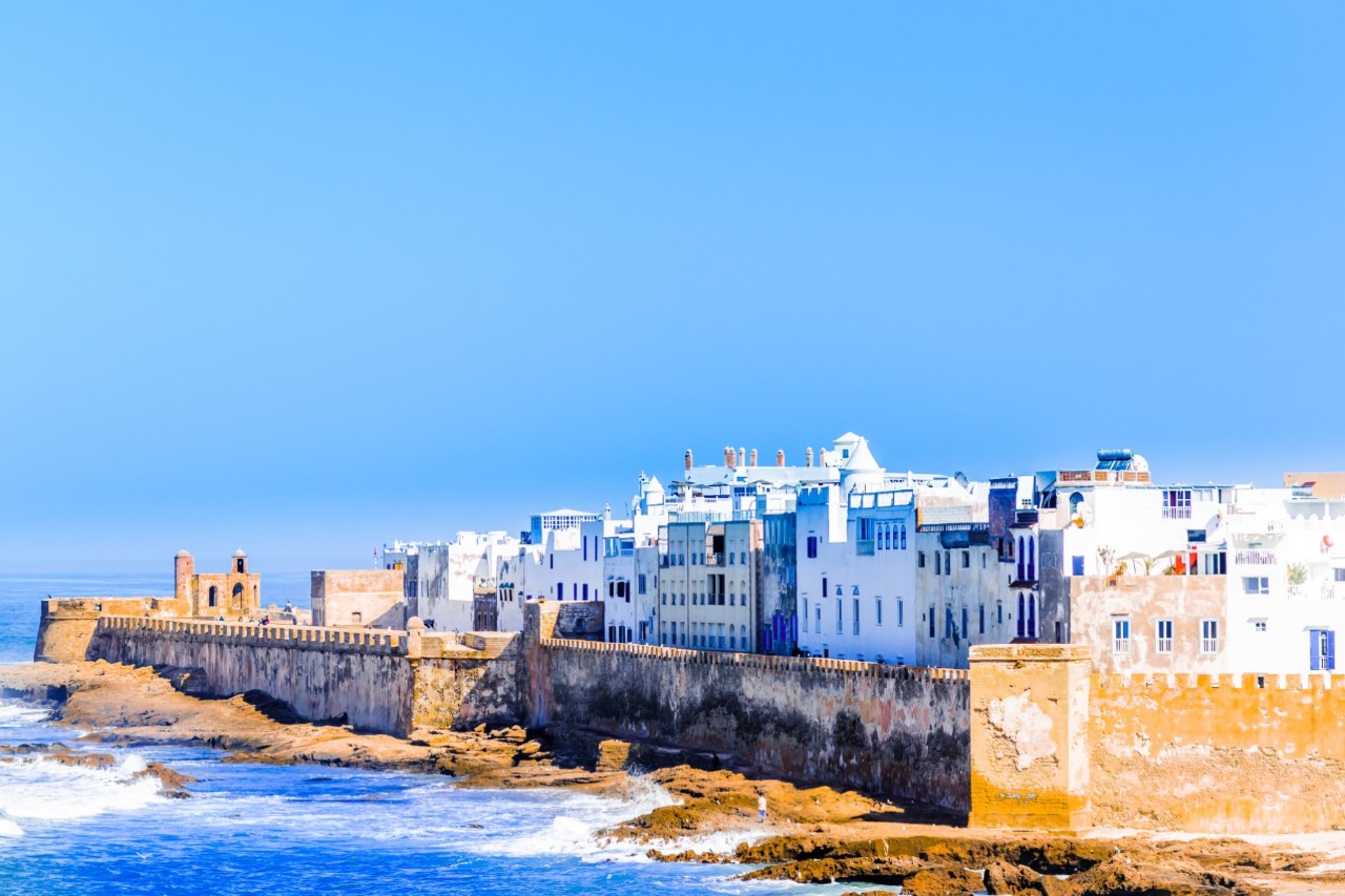 A coastal town with blue and white houses surrounded by a massive city wall. The city wall is topped with battlements. On the left, the sea rises up to the wall. The sky is blue.