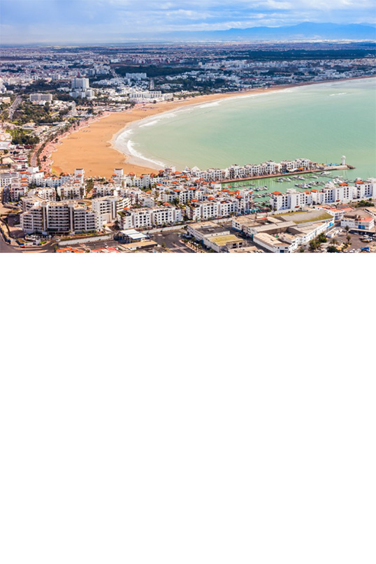 Aerial view of a large, modern coastal town. In the foreground, two rows of houses with multi-storey buildings jut out into the sea.