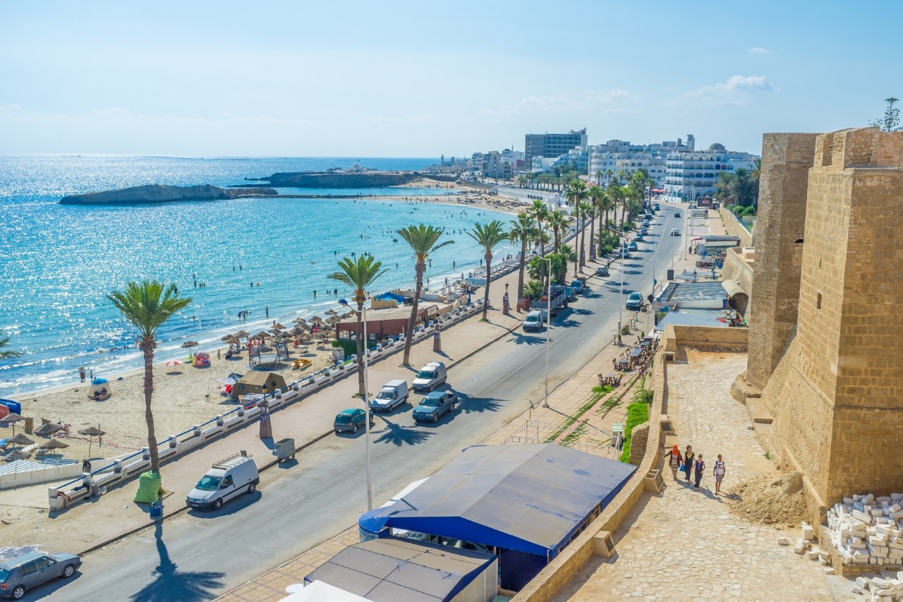 Coastal promenade. On the left side, the blue sea and a sandy beach with palm trees, parasols and bathers stretch out, while fortress walls can be seen on the right. A road with cars driving and parking, as well as palm trees, separates the promenade from the beach.