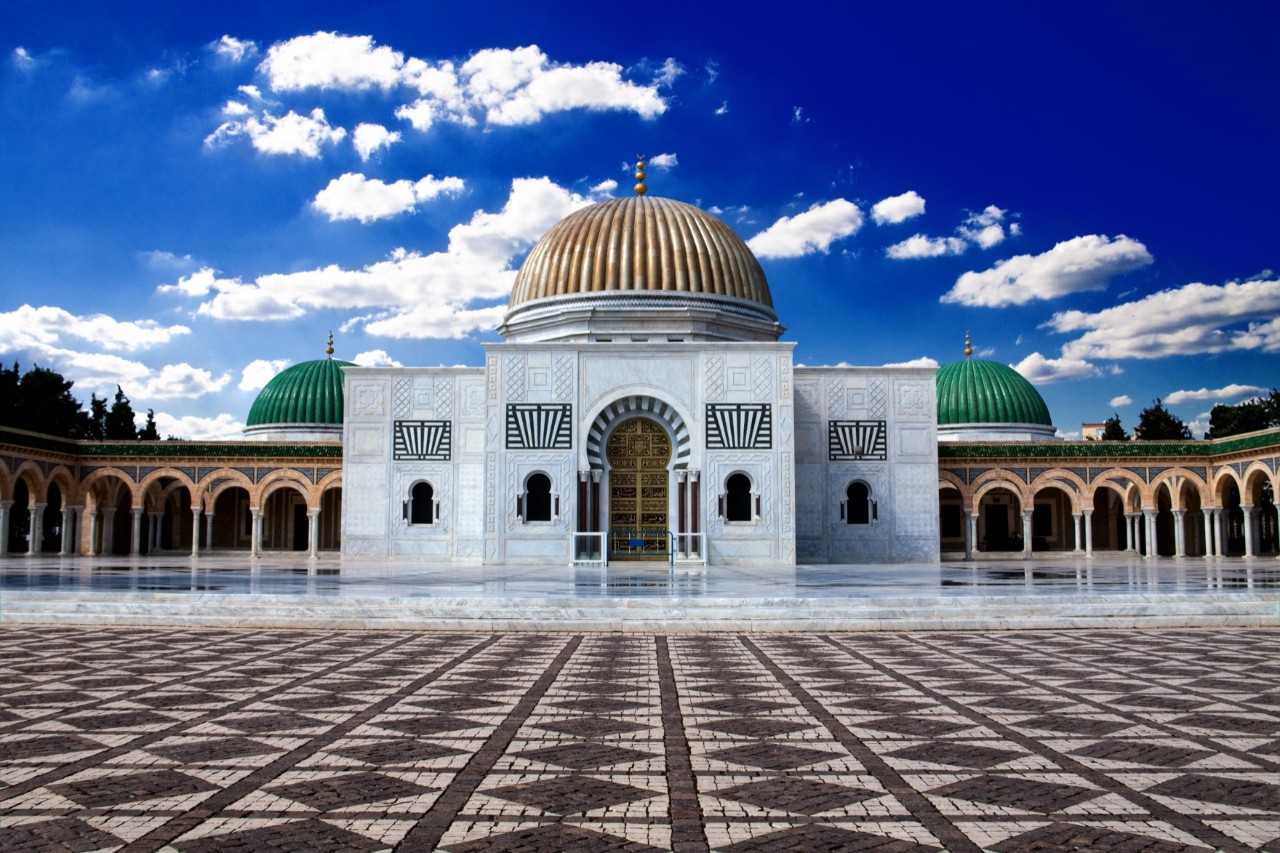 Magnificent white building with a golden dome in the centre and green domes on the sides. Arcades extend to the left and right of the building. The courtyard in front features a striking floor pattern.
