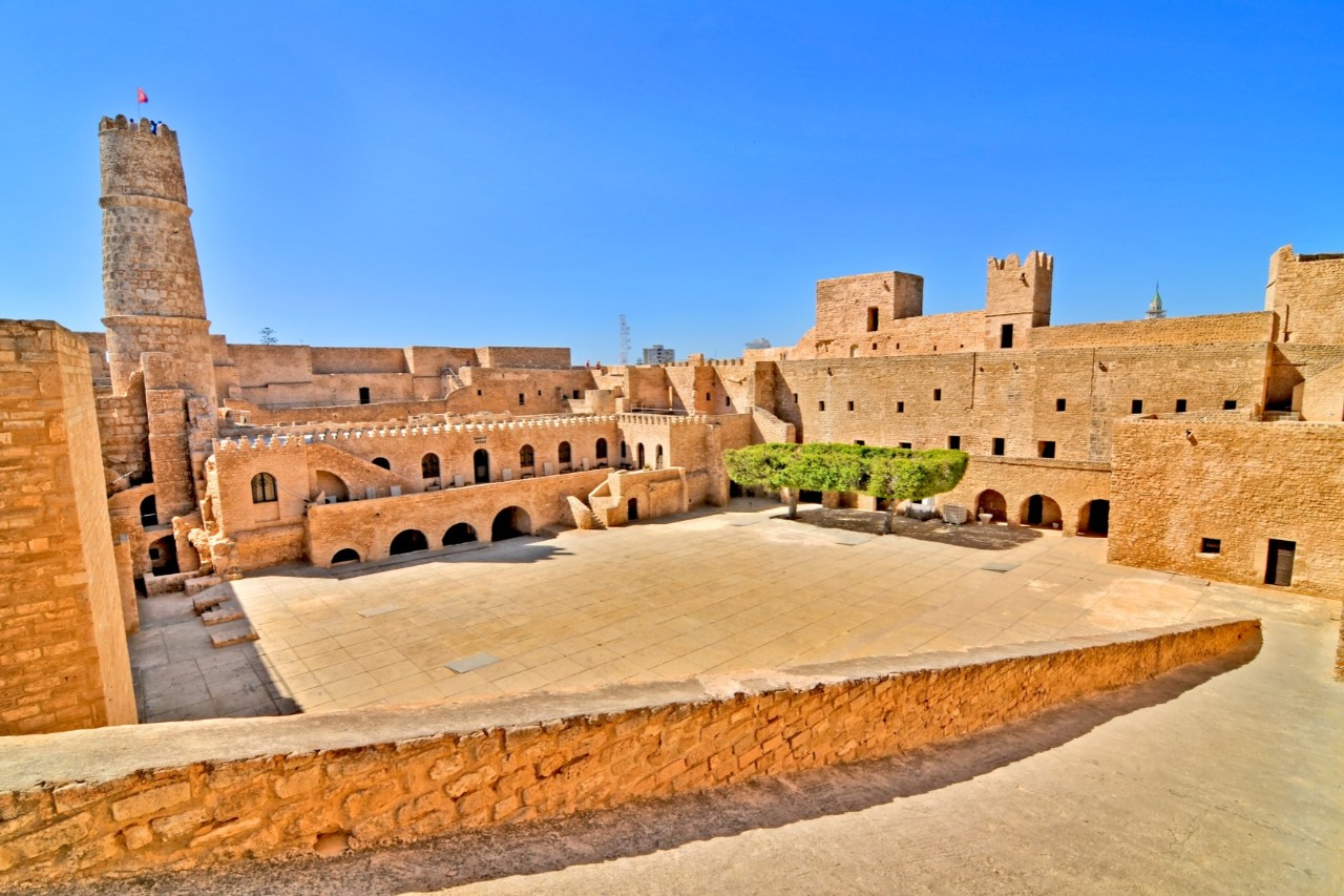 Inner courtyard of a fortress with ochre-sand-coloured stone architecture. The square is surrounded by massive walls, arcades, green trees and a tall tower with a flag flying on top.