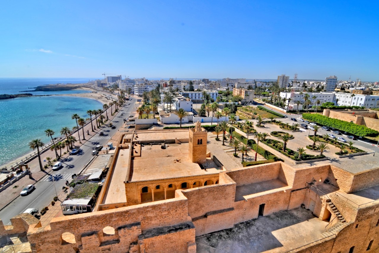 Bird's-eye view of an old fortress with sandstone architecture and a central tower. In the background, you can see a coastal promenade with palm trees, the blue sea and city buildings.