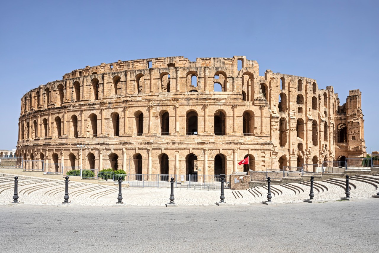 The picture shows a well-preserved, historic sandstone amphitheatre with several levels of arches and arched windows. The building is surrounded by a paved area with bollards.