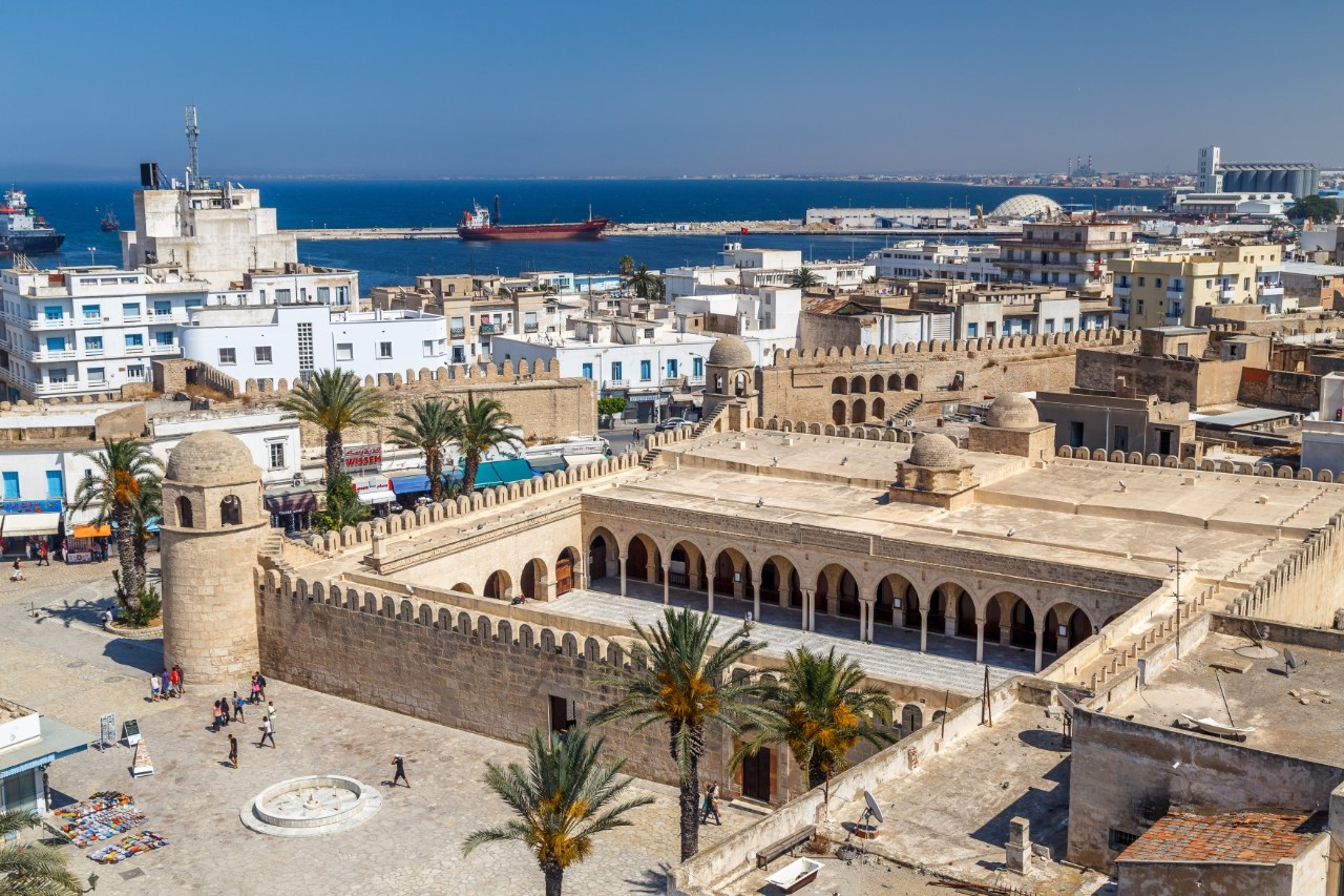 Cityscape with a sandstone fortress that includes arcades and towers and is surrounded by palm trees and a market square. In the background, the sea can be seen with ships in the harbour and modern buildings along the coastline.