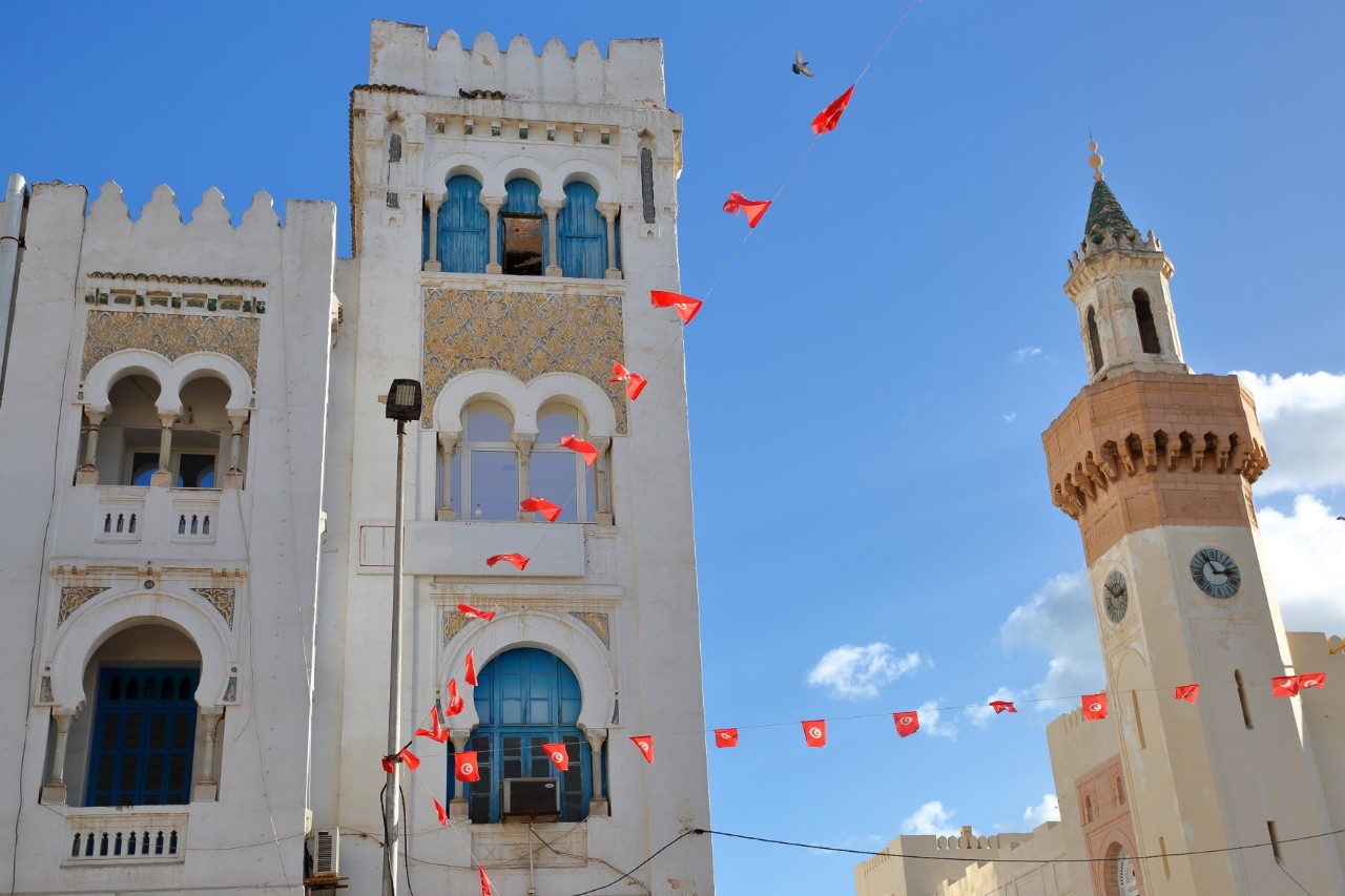 The picture shows older buildings with oriental arches and a detailed façade: on the left, a white house with blue windows and decorations, and on the right, a minaret with a clock and a spire. Red, rectangular pennants are stretched across the street.