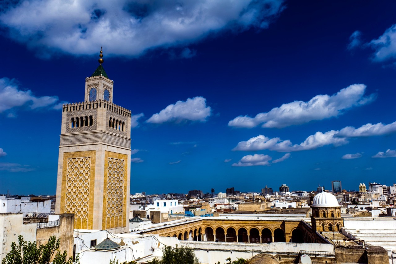 The picture shows a minaret decorated with geometric patterns against a deep blue sky with a scattering of white clouds, surrounded by a mixture of traditional and modern buildings.