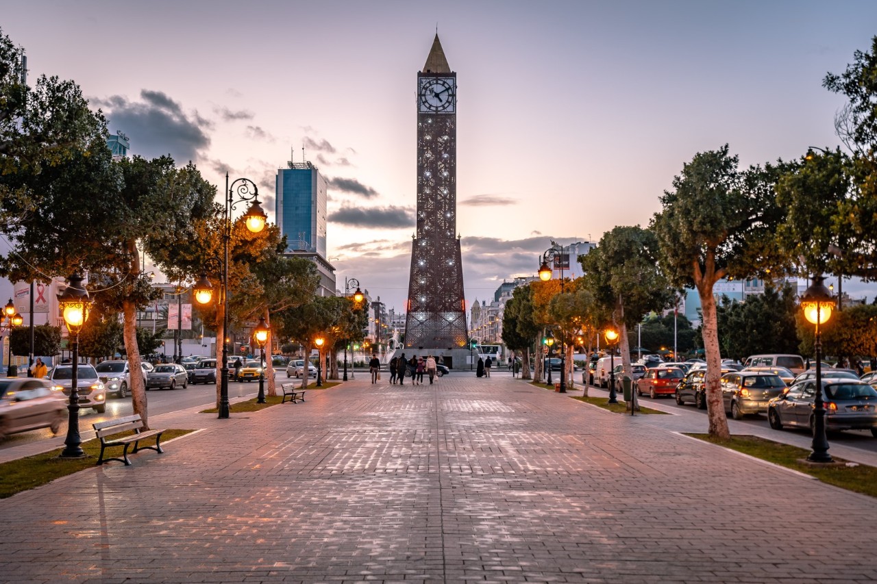 The picture shows a wide street with a pedestrian zone in the middle. There is a clock tower in the centre, flanked by trees and glowing lanterns on both sides along the street. People are walking, and cars are parked on the side of the road. It is dusk.
