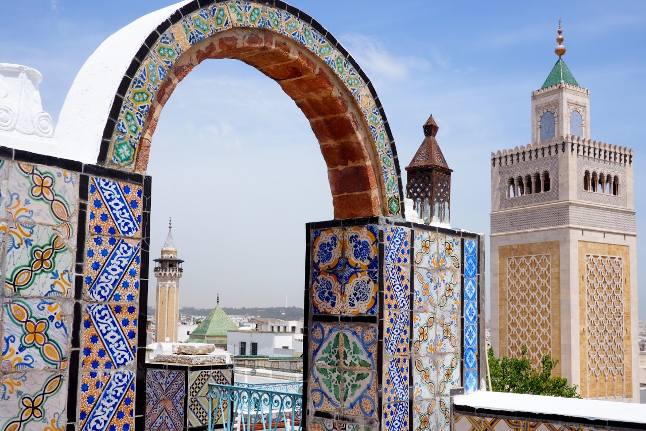 The picture shows traditional oriental architecture. In the foreground, there is an elaborately decorated archway with colourful, hand-painted tiles and arabesque patterns. Two minarets can be seen in the background.