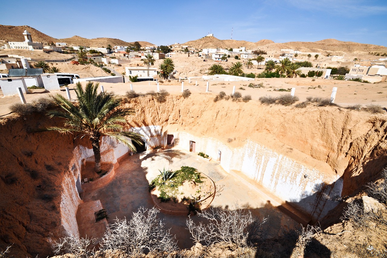 : Houses dug into the ground with a round courtyard and palm trees. In the background, a desert landscape stretches out with more palm trees and white houses under a blue sky.