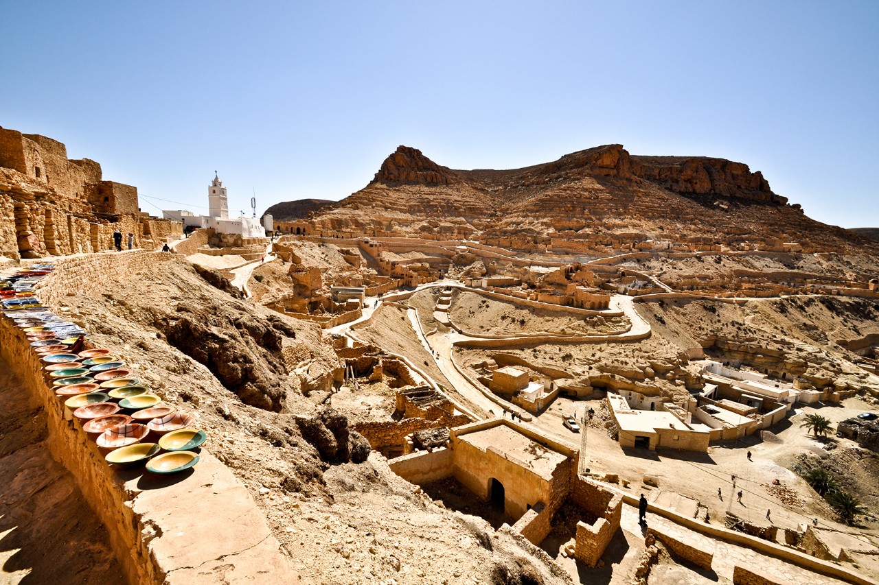 View of a mountain village with colorful ceramic bowls in the foreground, terraced buildings on the right, and a bright minaret in front of a striking desert mountain in the background.
