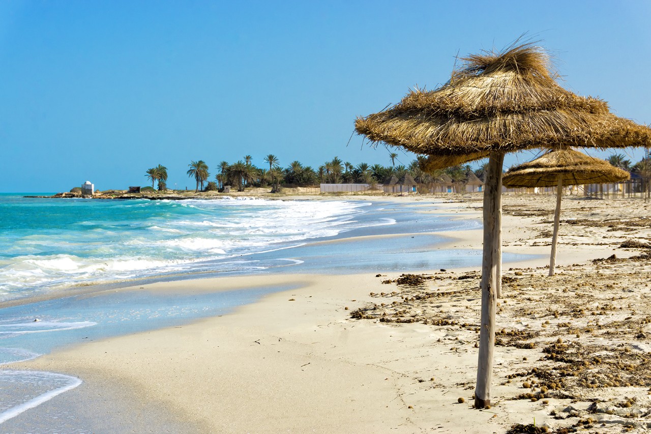 Wide sandy beach with palm trees, turquoise sea, and straw-roofed parasols. In the foreground, gentle waves roll onto the shore.