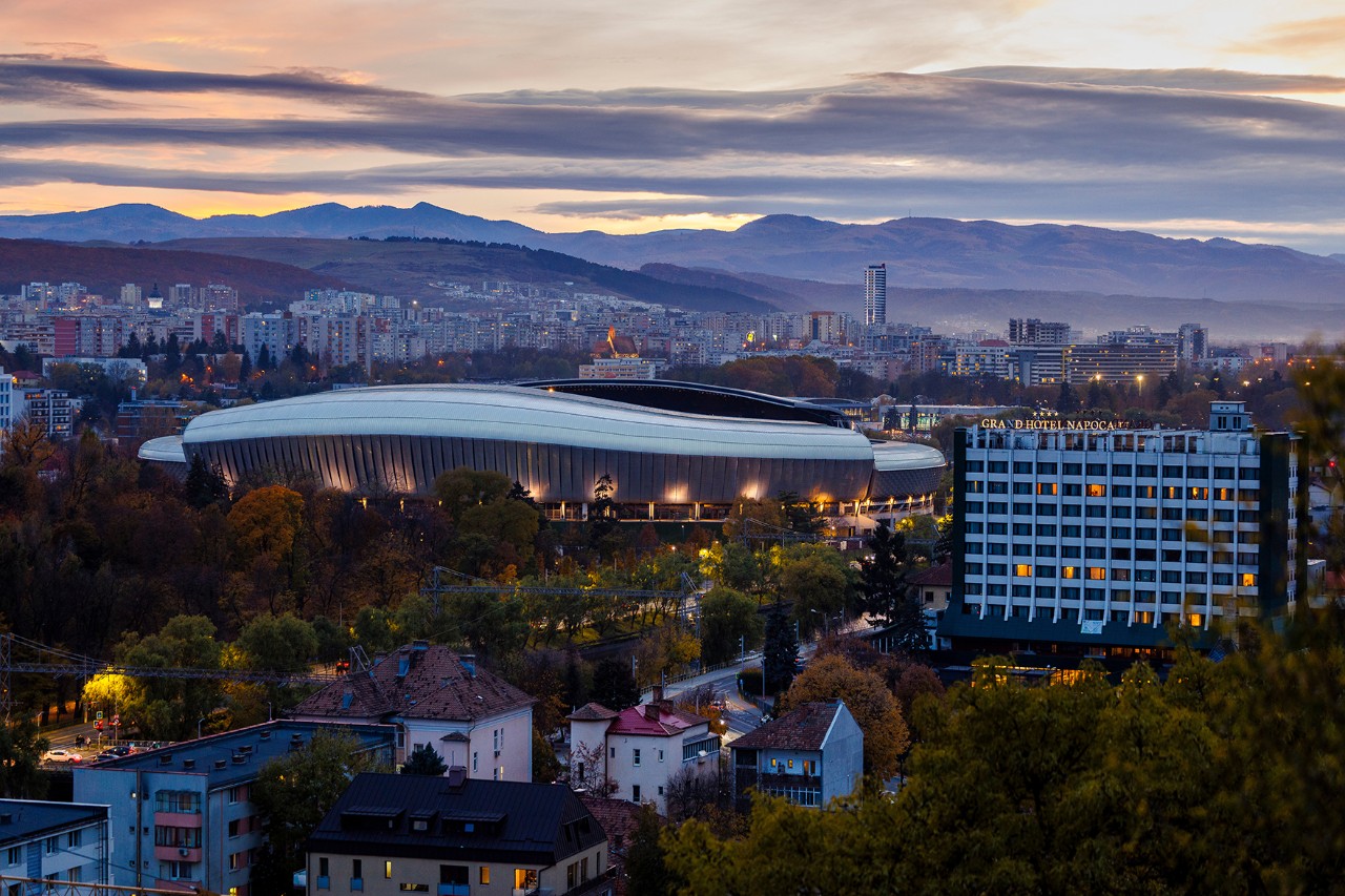 The image shows a sports stadium surrounded by trees in the centre of a large city, with a mountain range in the background. The lighting is dim.