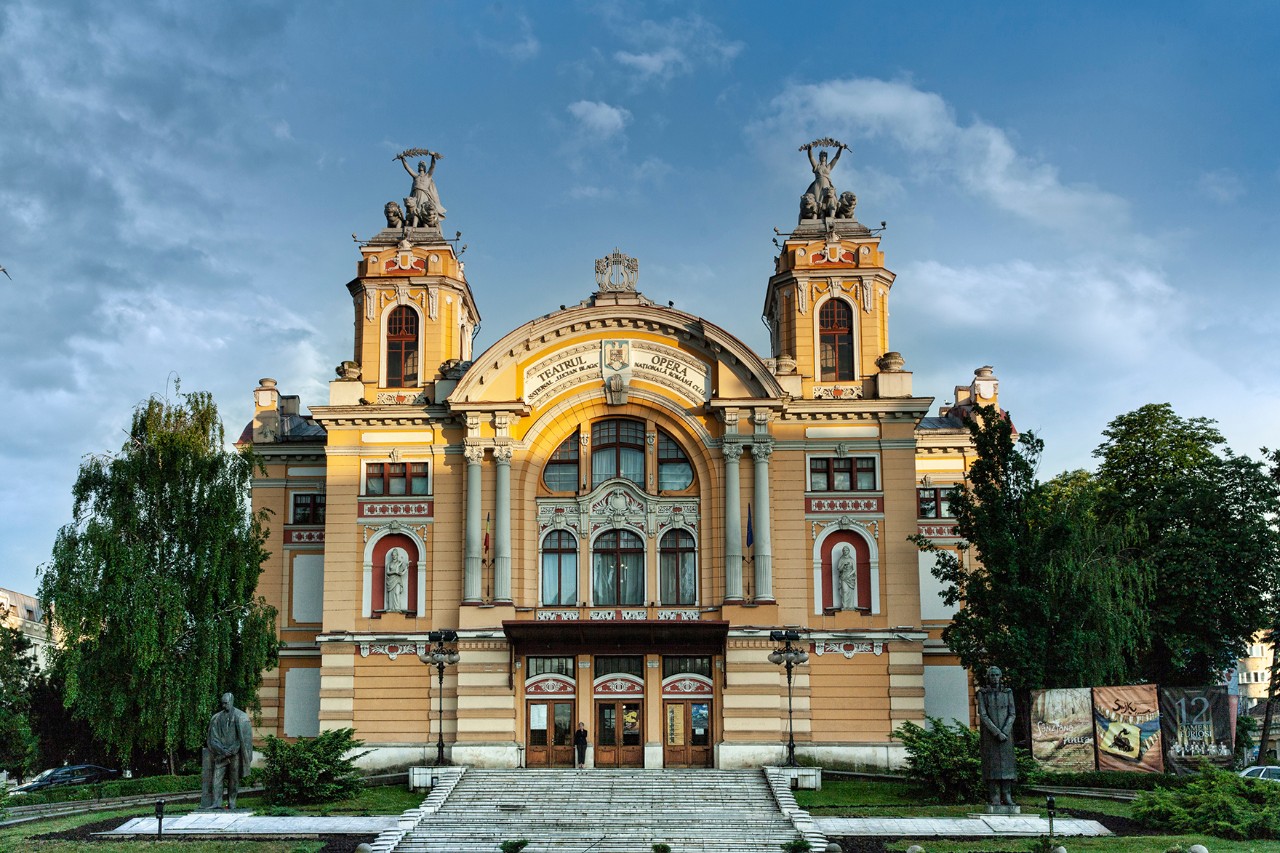 In the picture, you can see the front of a building. The yellow façade is decorated with columns and statues. Turrets and arches emphasise the neo-Baroque style.
