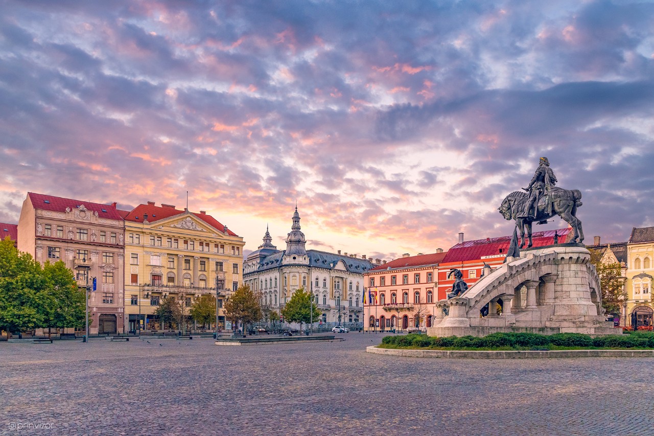 The picture shows a paved square with an equestrian statue. Various old buildings with colourful facades and trees frame the square.