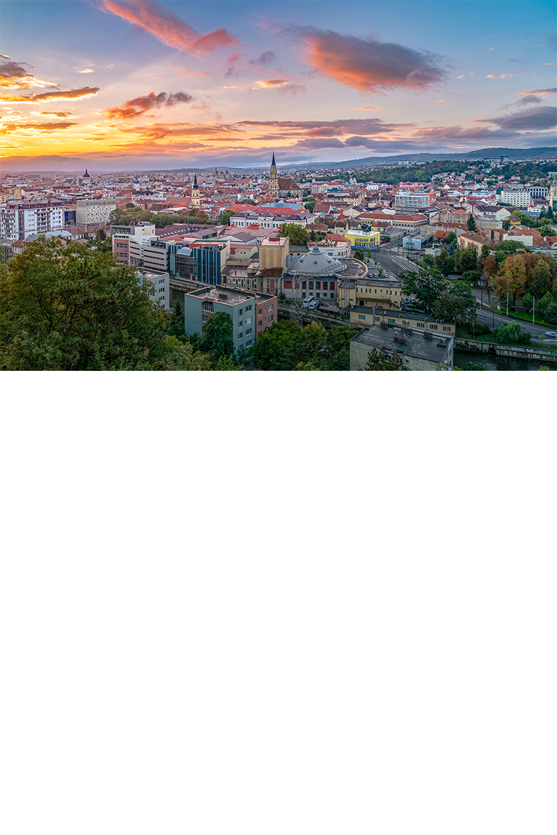 The image shows a large city in the light of the setting sun, with several church towers rising above other buildings and green hills visible in the background.