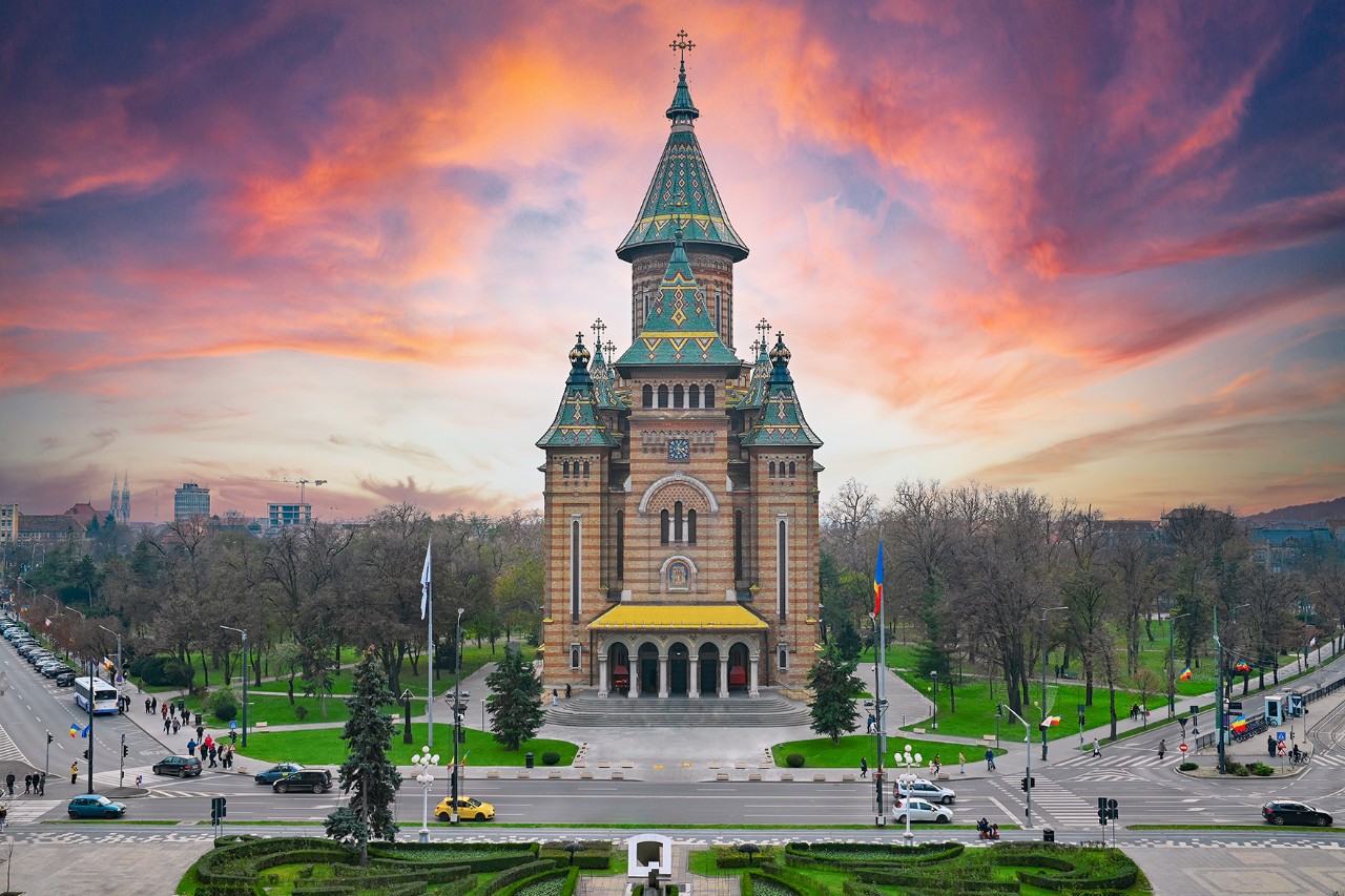 Large church with six green spires in front of a park and a busy road, dramatic sunset in the sky.