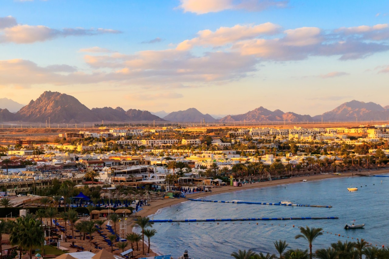 Coastal town with white buildings and palm trees, a beach with sunbeds and the calm sea in the foreground. In the background, impressive mountains tower up.