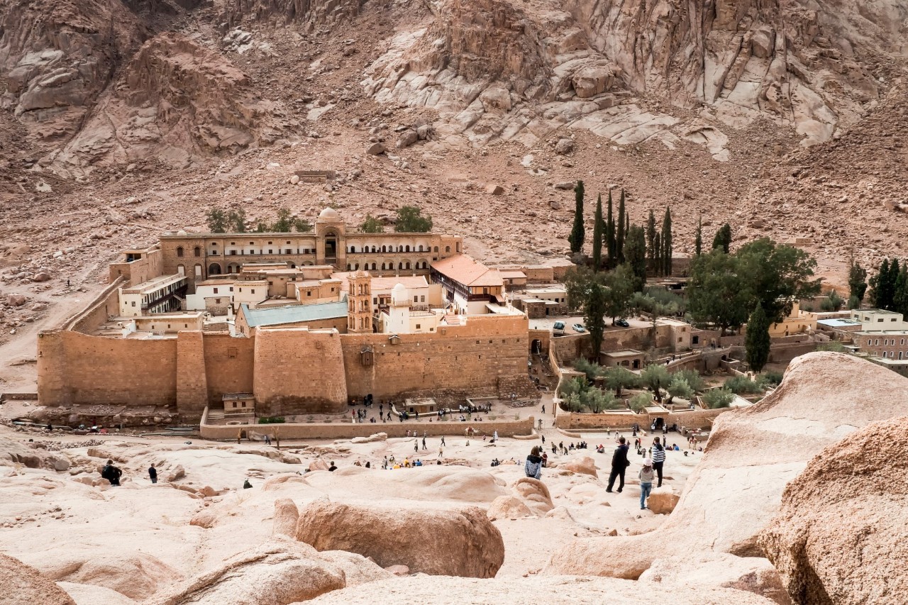 Old monastery surrounded by rocky mountains. In the foreground, visitors can be seen on rocky terrain. The monastery has massive sandstone walls and cypress trees that frame the building.