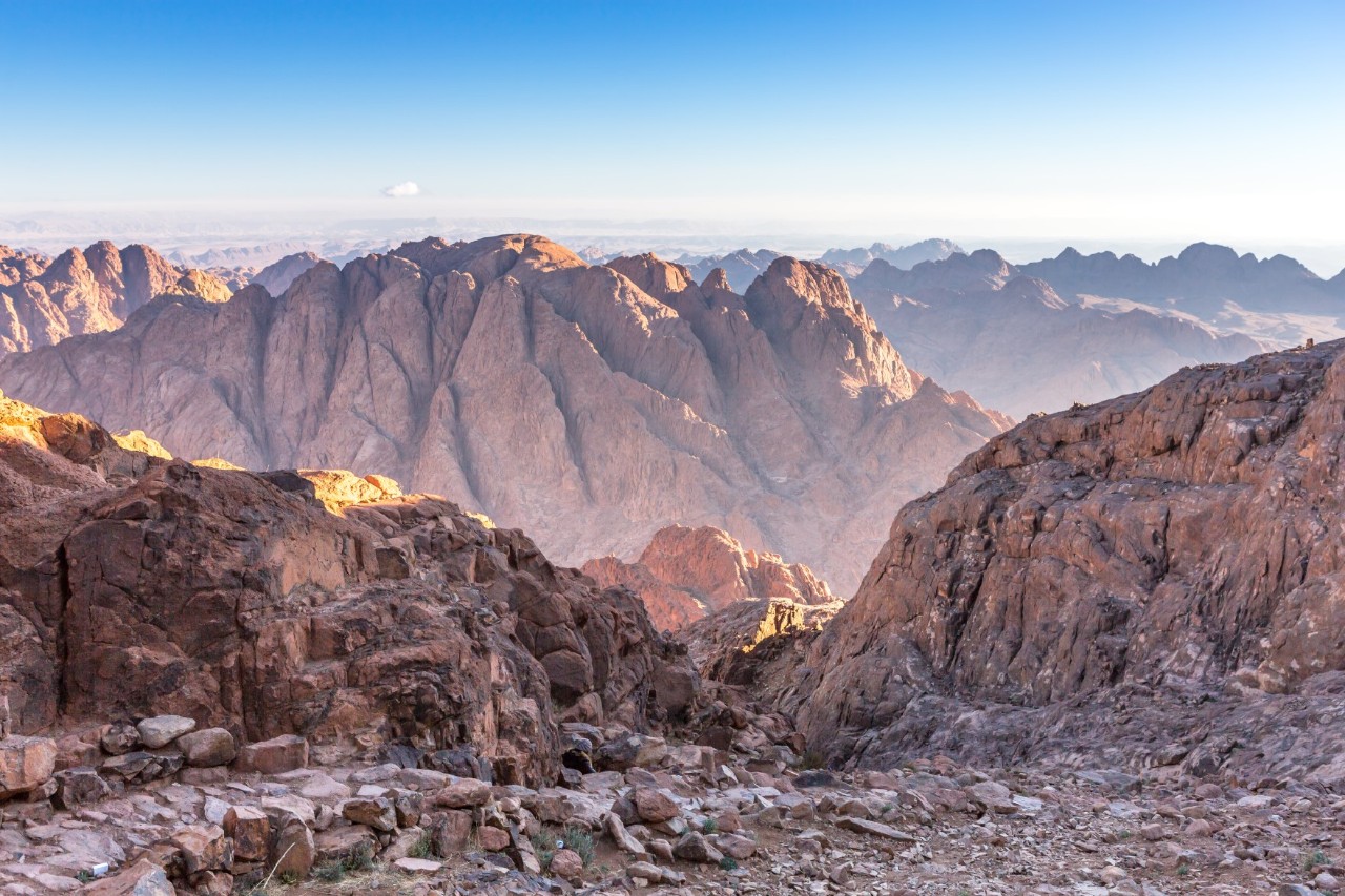 Reddish mountain landscape with high peaks bathed in warm sunlight. Rocky paths can be seen in the foreground.