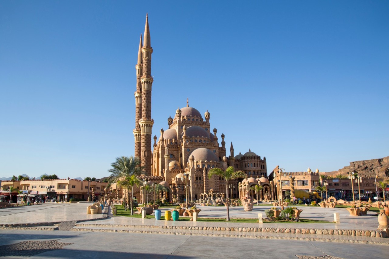 Impressive mosque with several domes, ornate details and two tall minarets. In the foreground, there is a paved square, palm trees and decorative elements.