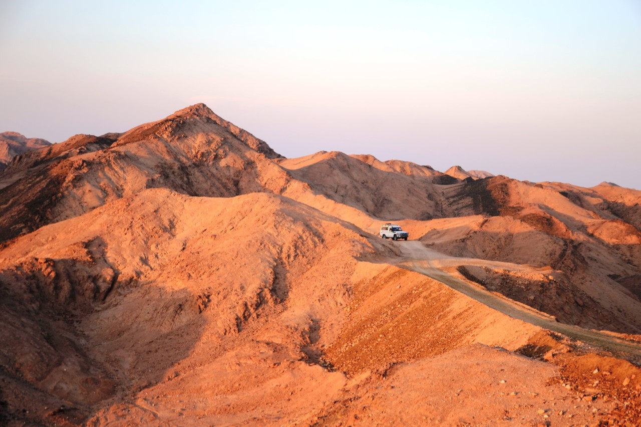 A barren desert landscape with brownish-red hills. An off-road vehicle is driving along a narrow, unpaved road that winds through the hills.