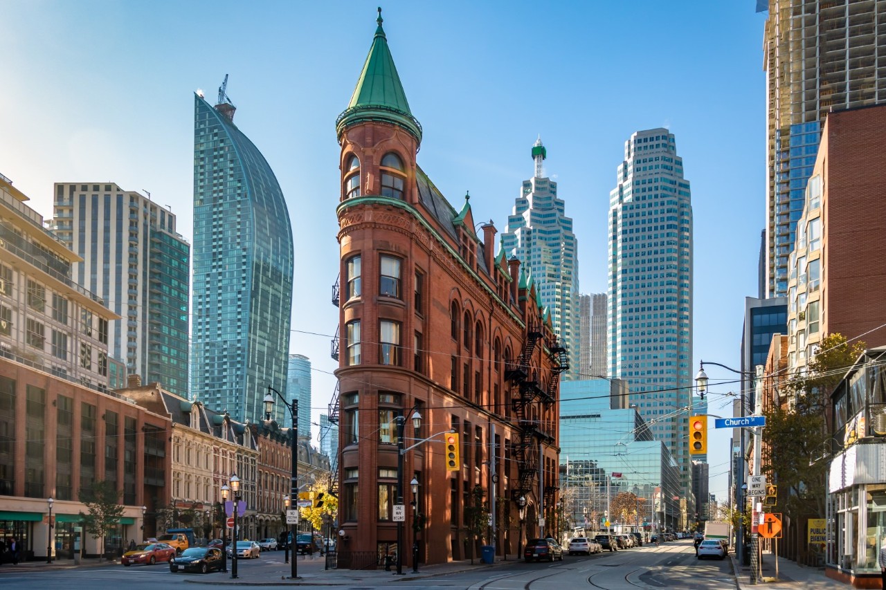 A historic red brick building with green copper towers in a triangular shape. Modern skyscrapers rise up in the background. Traffic lights, cars and rails can be seen in the foreground.