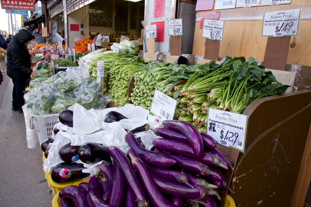 A market stall with fresh vegetables in an urban environment. Purple aubergines are lying in the foreground, behind them are various green vegetables such as bok choy and broccoli. The signs show the prices and labels in English and Chinese characters. On the left, a person can be seen looking at the offer. The market stall looks well stocked and offers a wide selection of fresh vegetables.