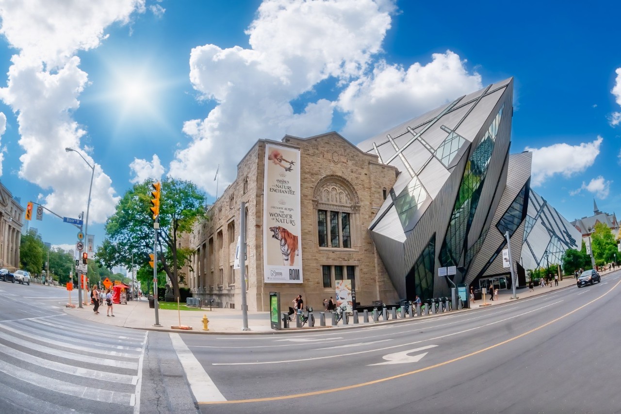 A striking museum building, consisting of a historic stone building and a modern glass and metal extension in an angular style. In the foreground is an intersection with a zebra crossing and pedestrians.