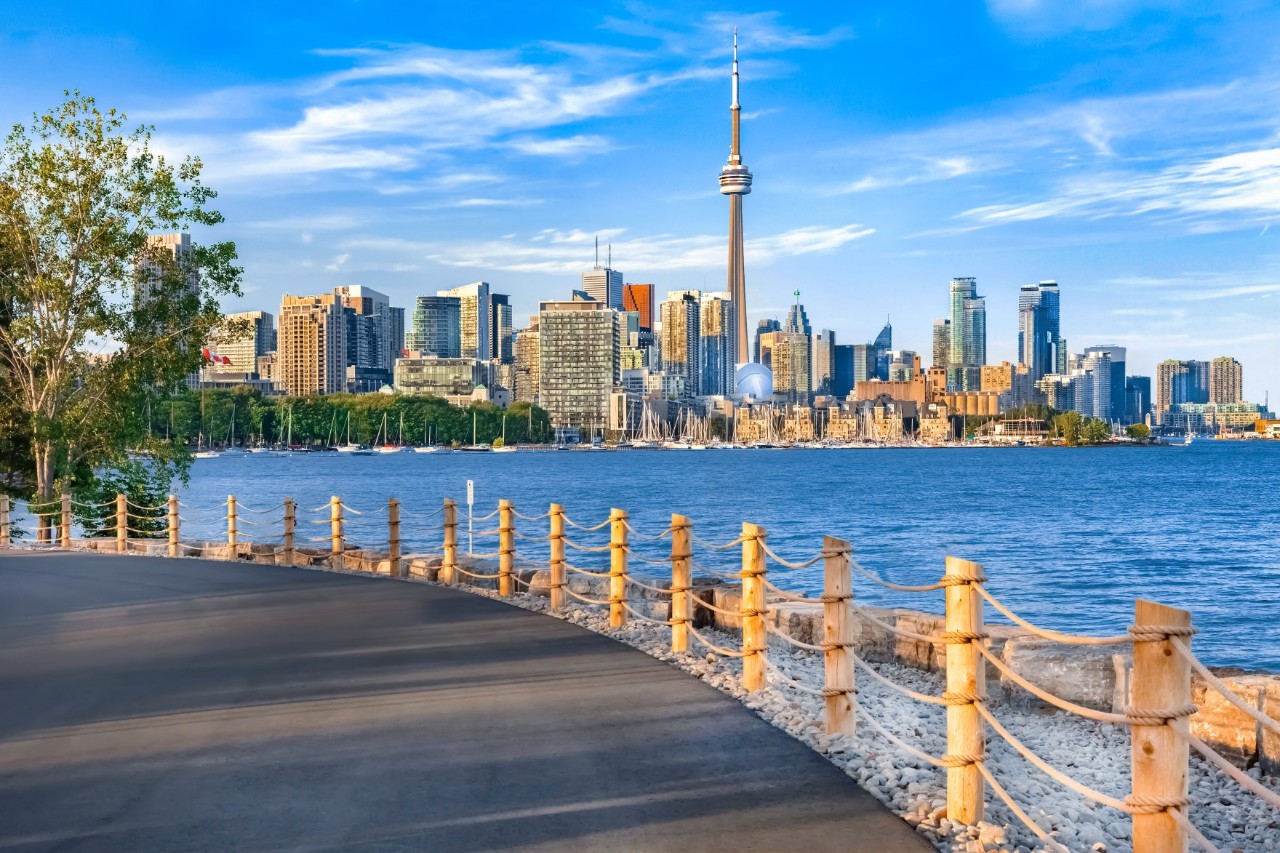 A skyline with a television tower in the centre, taken from a lakeside path along a large lake. In the foreground, there is a paved path with wooden railings.