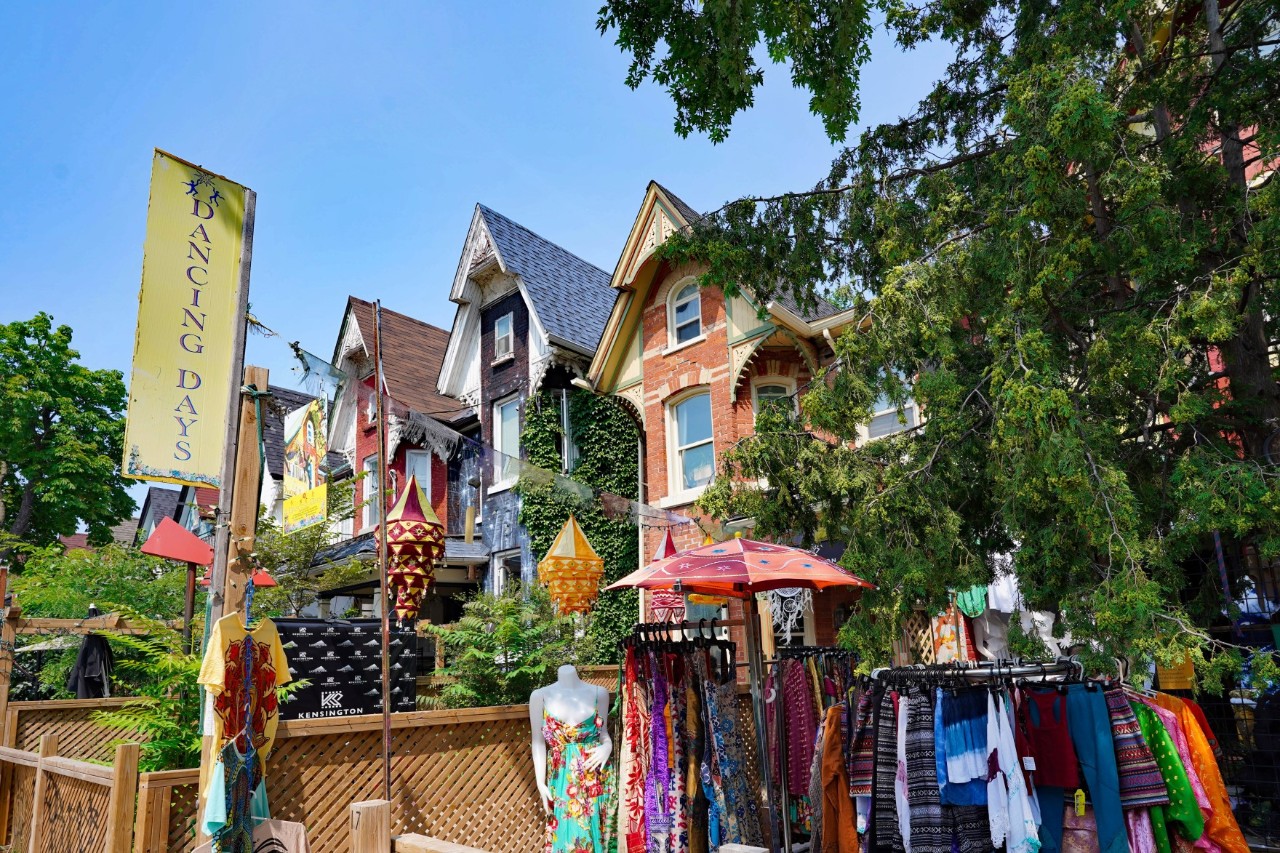 A street with Victorian houses, some of which are covered with ivy and surrounded by trees. In the foreground, there are stalls with colourful clothing and a sign saying ‘Dancing Days’.