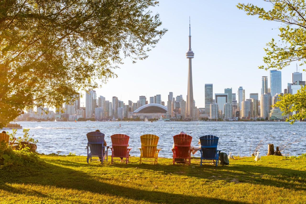 Coloured Adirondack chairs are placed in a meadow by the water, with a view of the Toronto skyline, including the striking CN Tower. The calm water reflects the sunlight, while trees form the picture frame on the left and right. The scene is idyllic and invites you to relax, while the city in the background adds a sense of vibrancy.