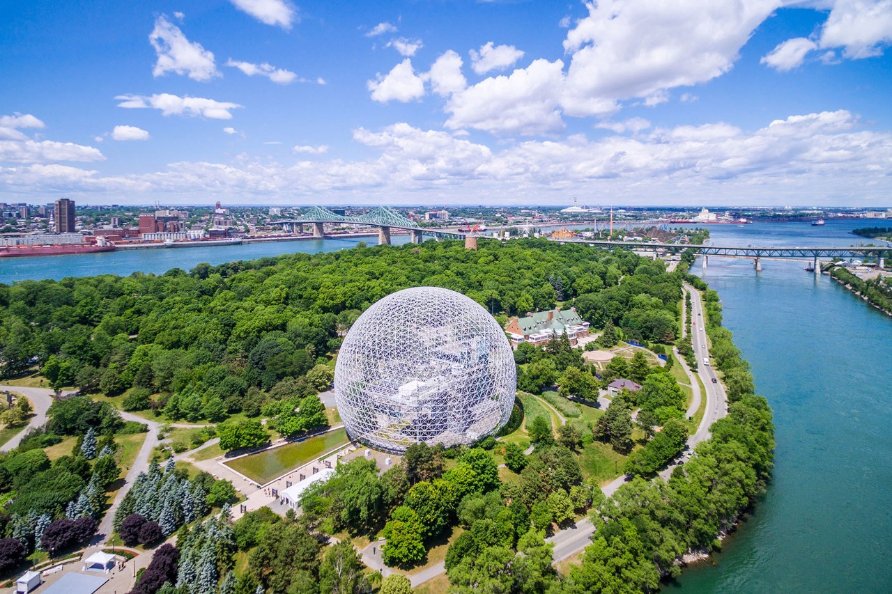 Aerial view of a green island with a striking spherical geodesic dome in the centre, nestled in a park landscape. The island is surrounded by water, connected by bridges, with an urban skyline in the background.