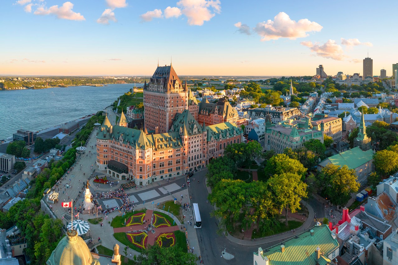 Imposing building with many towers and bay windows in the French Renaissance style, surrounded by other smaller houses, streets and trees. A wide river flows to the left.