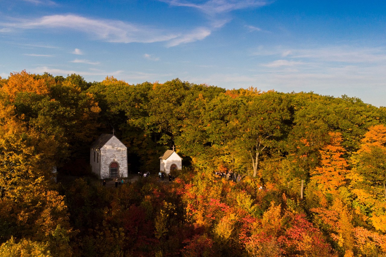 Aerial view of a national park with two small chapels surrounded by dense forest in autumn colours under a blue sky.