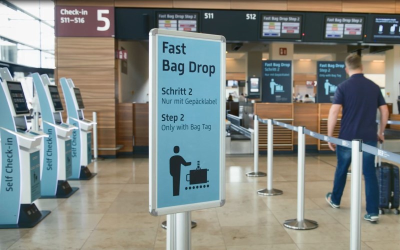 Blick auf ein Schild mit Fast Bag Drop INformationen. Im Hintergrund Automaten und Schalter für die Gepäckabgabe.
