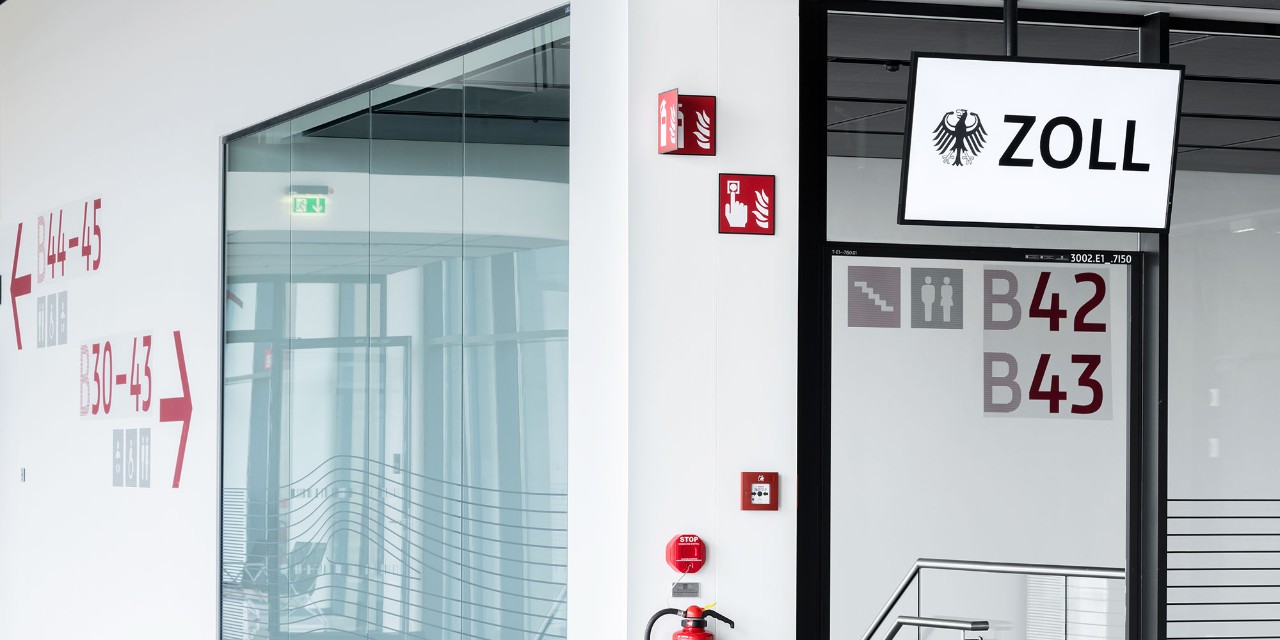 Glass wall with a monitor displaying the word 'Zoll', customs.