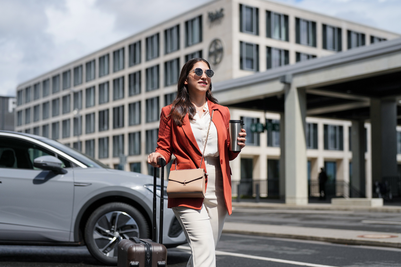 Woman in business outfit with orange blazer, light-coloured trousers and sunglasses, walking past a modern building and parked car, pulling a trolley case and holding a coffee-to-go.