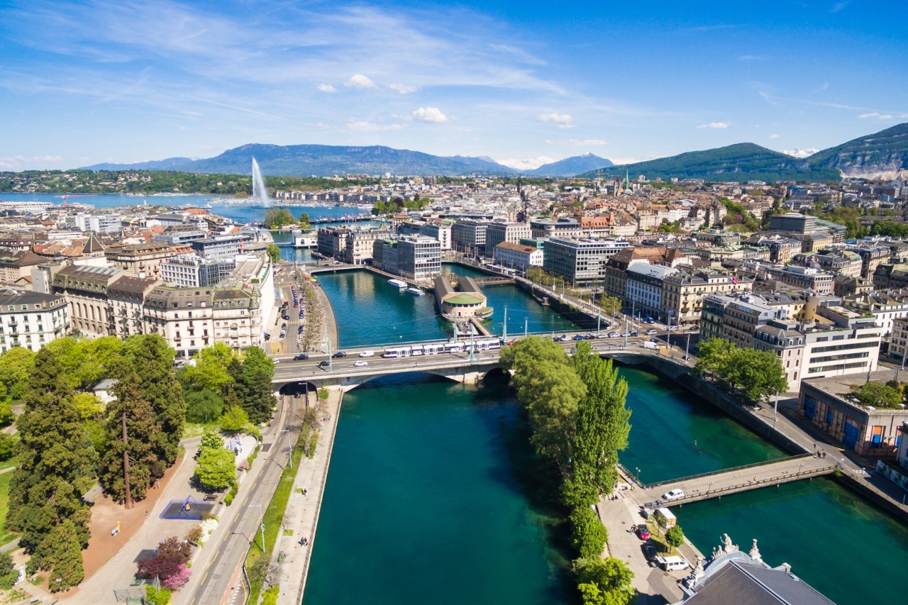 View from above of a green-blue river running through a city and flowing into a lake. Several bridges cross the river. In between there are green island areas. Streets run along both shores of the river and next to them are multi-storey buildings. A mountain panorama can be seen in the background of the picture.