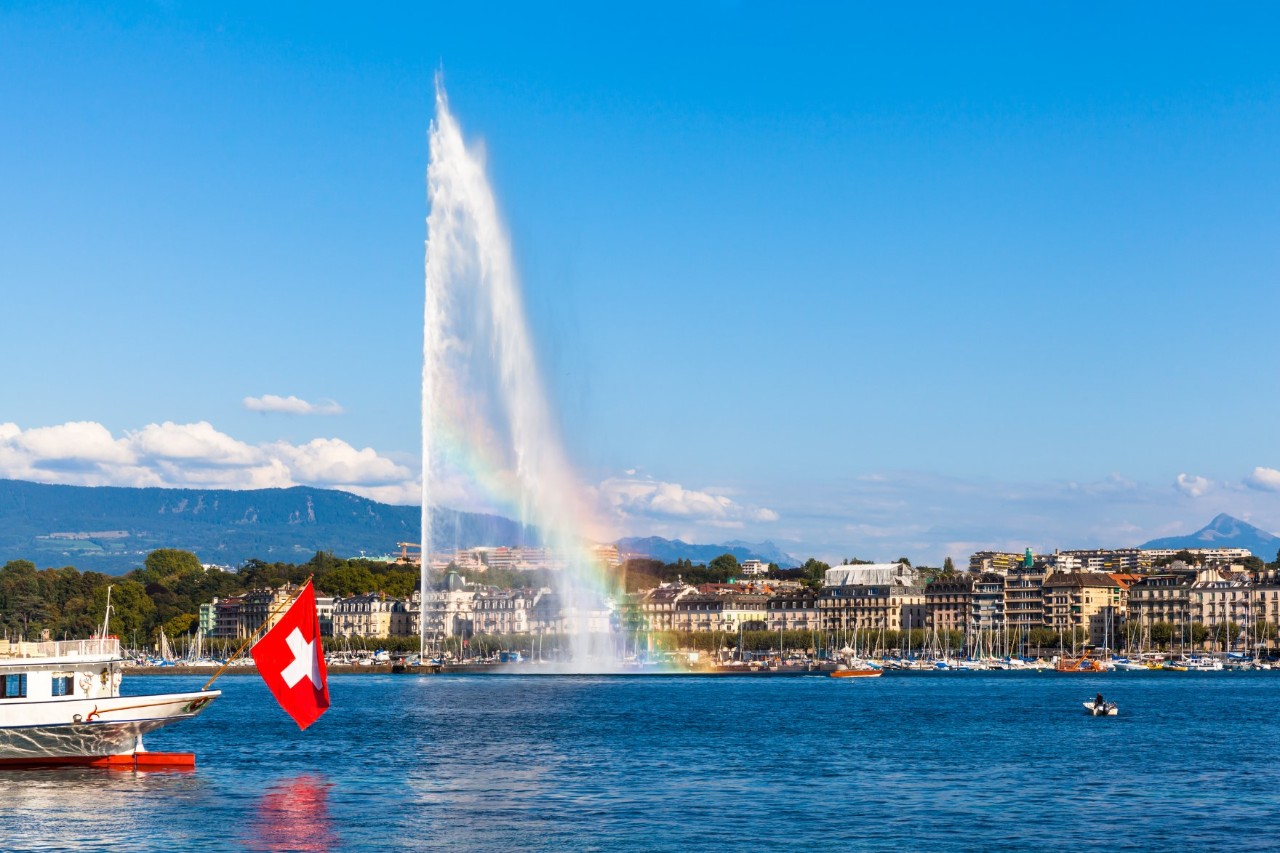 A water fountain rising into the air with a rainbow in the lake; in the background is a lakeside promenade with buildings. A small white boat with a Swiss flag, a white cross on a red background, is moored at the front left edge of the picture.
