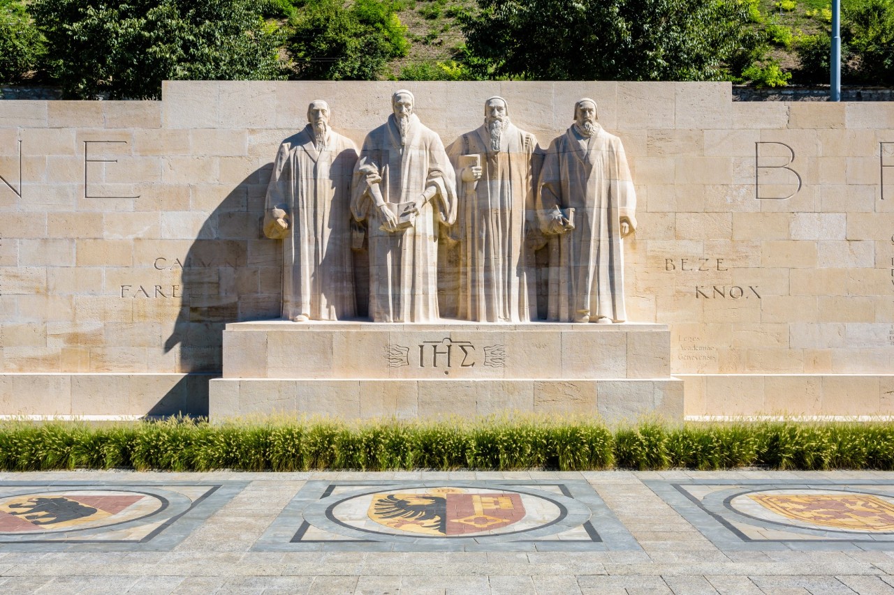 Four sandstone sculptures showing men in long robes on a pedestal in front of a wall. Three large coats of arms are set into a stone floor in front of the statues. The wall is lined with trees.