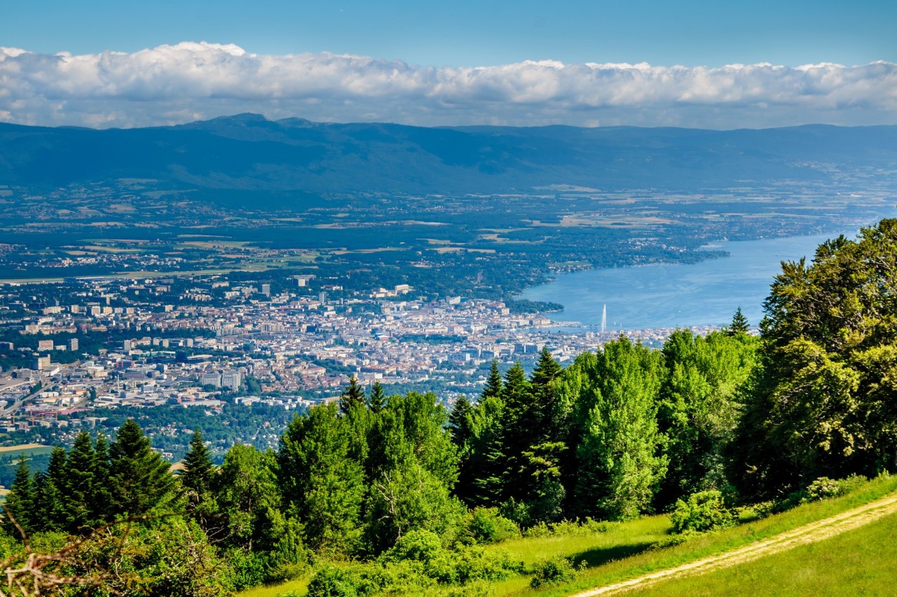 A distance from a tree-covered mountain to a far-away city on a lake. The mountains, and the clouds above them, can be seen in the background.