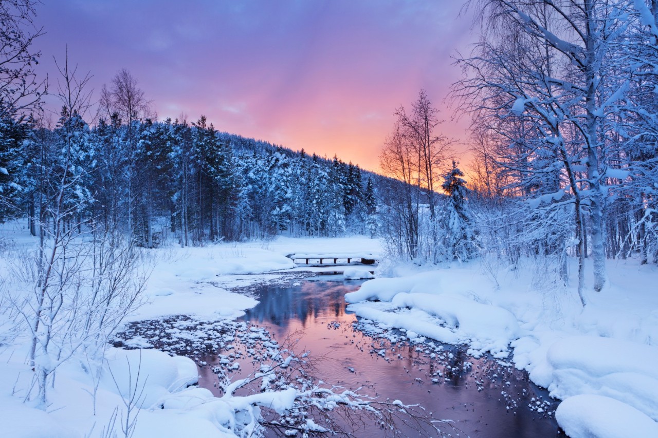 A snowy winter landscape with a river, snow-covered trees and a small wooden bridge in the light of the setting sun.