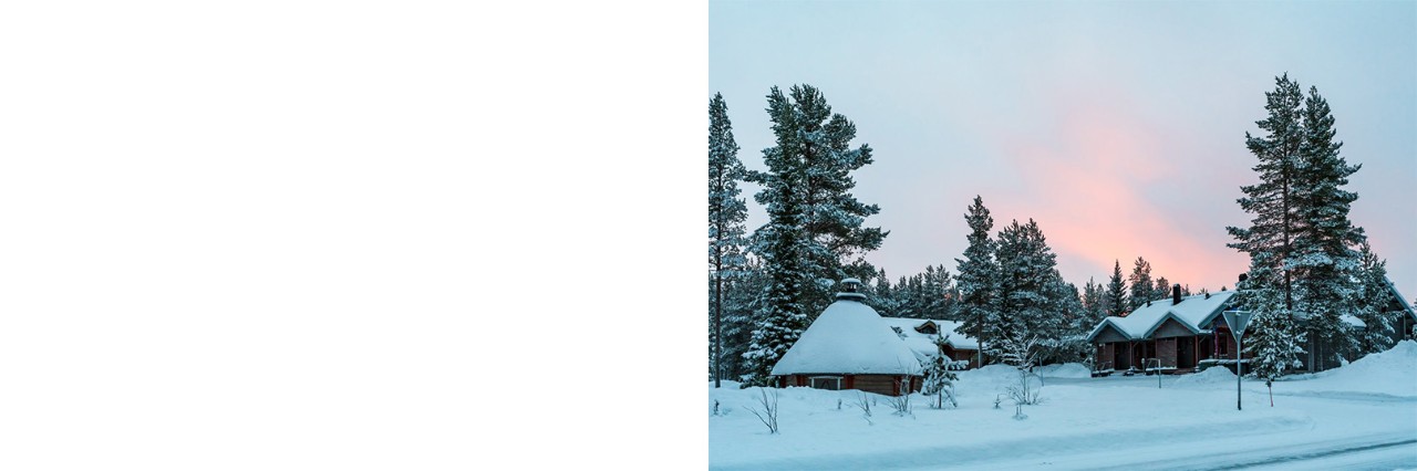 Snow-covered wooden houses in a winter forest with a partly pink sky and snow-covered trees. 