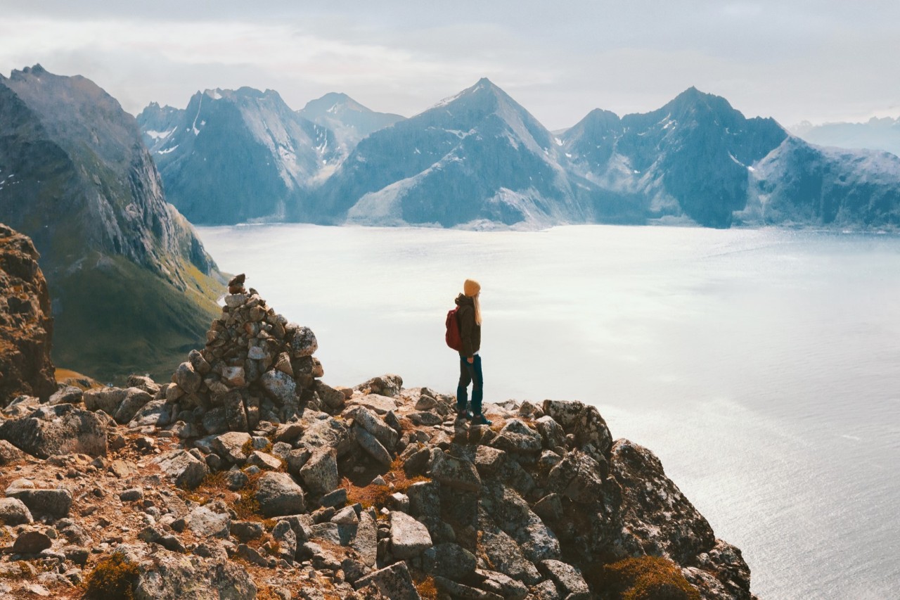 Hiker stands on rocky ledge overlooking fjord-like mountain landscape.
