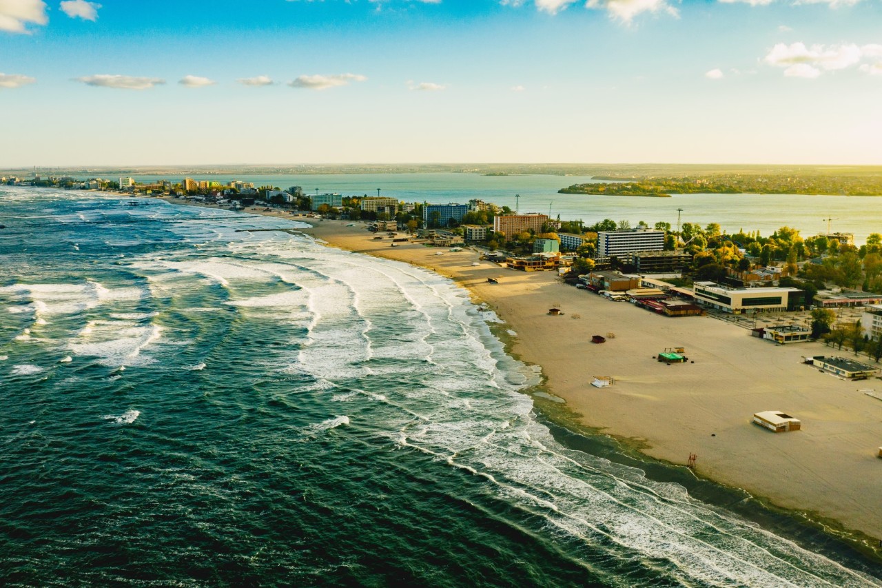 Buildings on a narrow headland leading into the sea with waves and a wide sandy beach. 