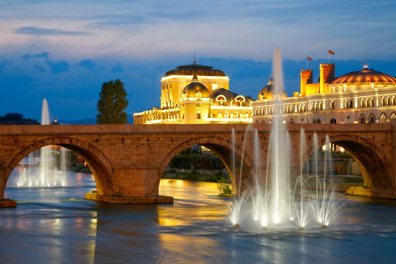 A bridge with arches over a river, in which several water fountains can be seen, at twilight. On the right the picture are striking, illuminated buildings.