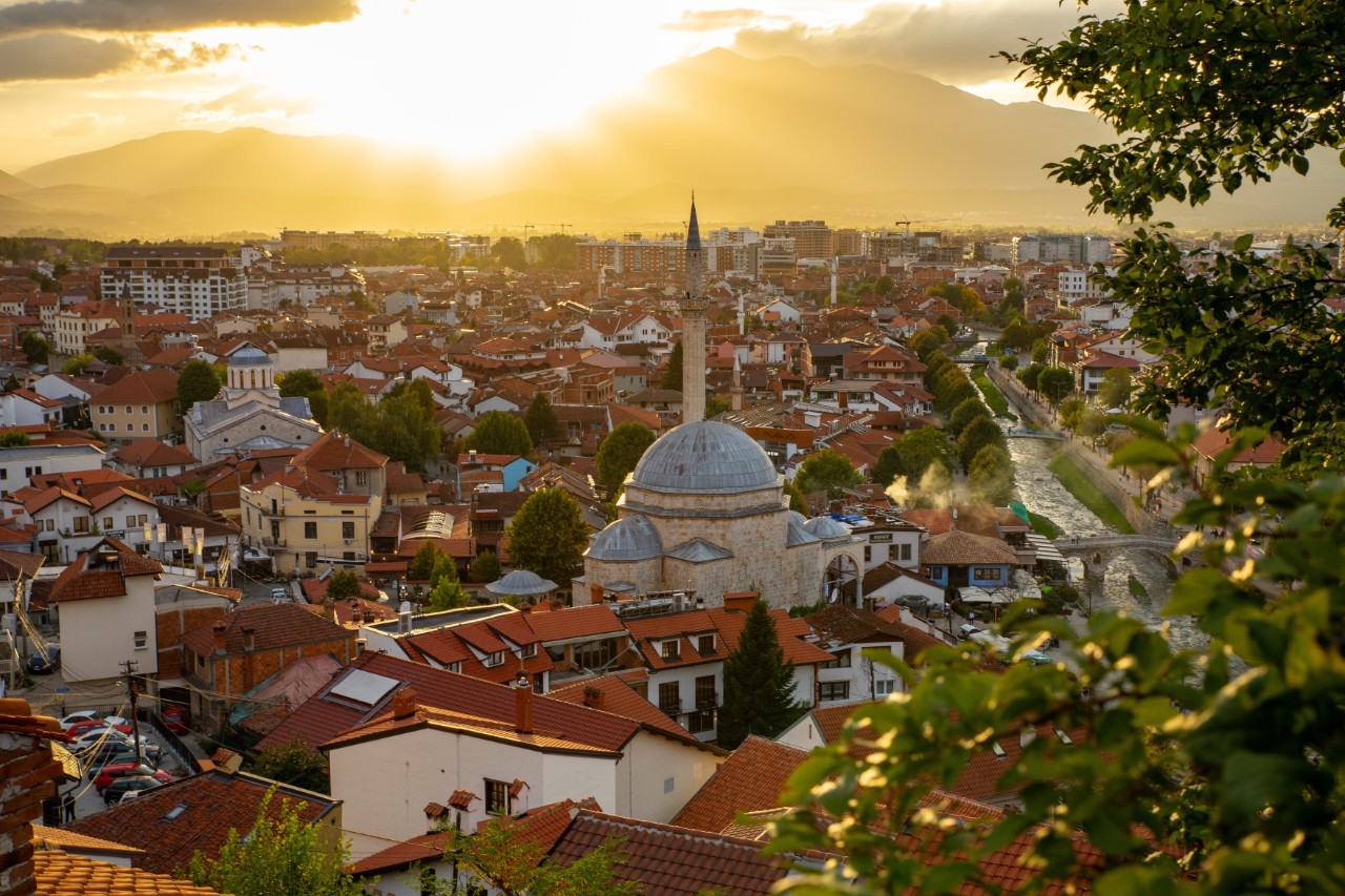 View from above of a city with numerous red tiled roofs and a mosque with a large dome and minaret in the centre. Green branches protrude into the picture in the foreground. The sun is shining on the horizon just before sunset, illuminating the city with warm light.