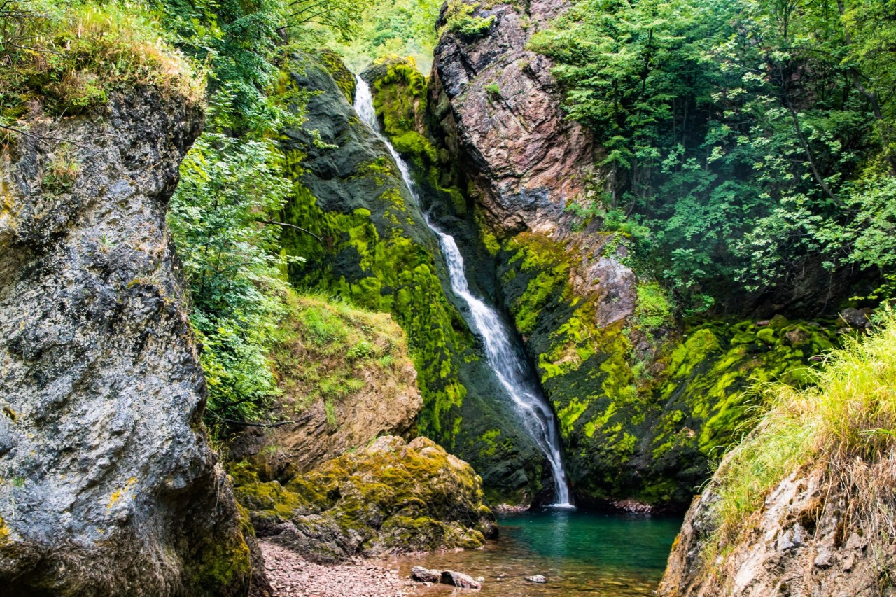 Lush, green, steep rocky landscape through which a high, narrow waterfall leads into a small, green lake.