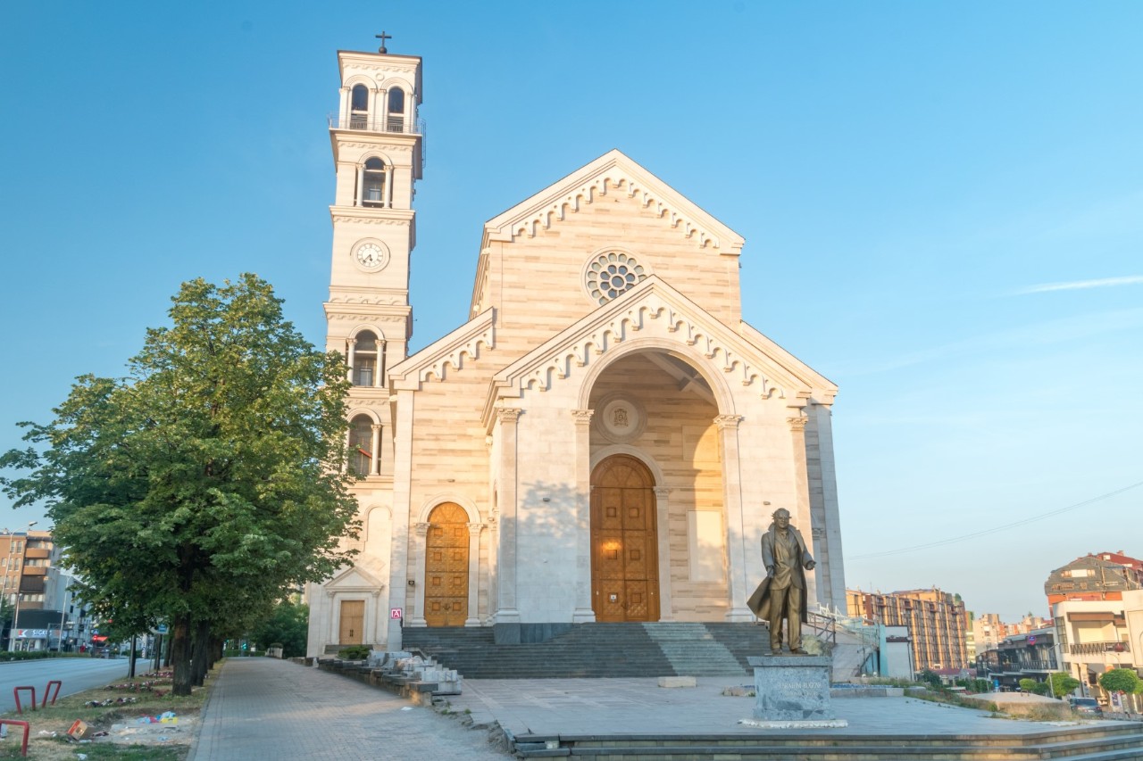 Large, bright church with a pointed roof and a high, arched entrance, which is accessed via a wide staircase. To the left of the nave is a large, square church tower with several arched windows and a clock. There is a statue in front of the church. To the left of it stands a large tree, while on the right side there are several houses lined up in a row.
