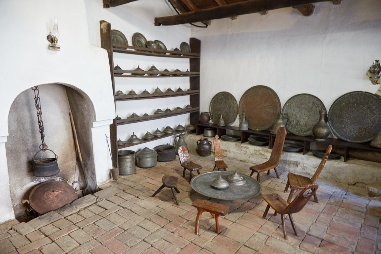 Old living room with several chairs around a round table on a brick floor. Behind it, an open fireplace is visible. Numerous cast-iron pans and pots can be seen on the table and on a shelf on the wall.
